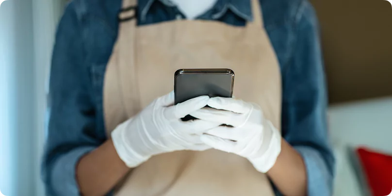 Camp cleaning staff in white gloves and beige apron using smartphone for service coordination at a resource sector accommodation facility.