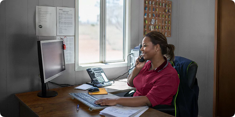 Smiling female staff member in red polo shirt answering phone while working at computer desk in workforce accommodation office.