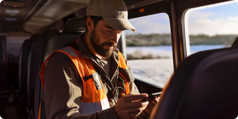 Bus-in bus-out worker in orange safety vest using mobile device while seated in a vehicle with snowy landscape visible through window.