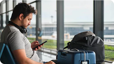 Man checking mobile phone while seated in airport waiting area with luggage including blue suitcase and backpack.