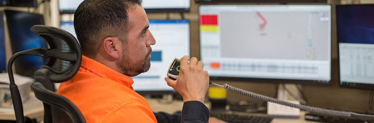 FIFO worker holds a dispatch device while working at a desk with multiple computers.