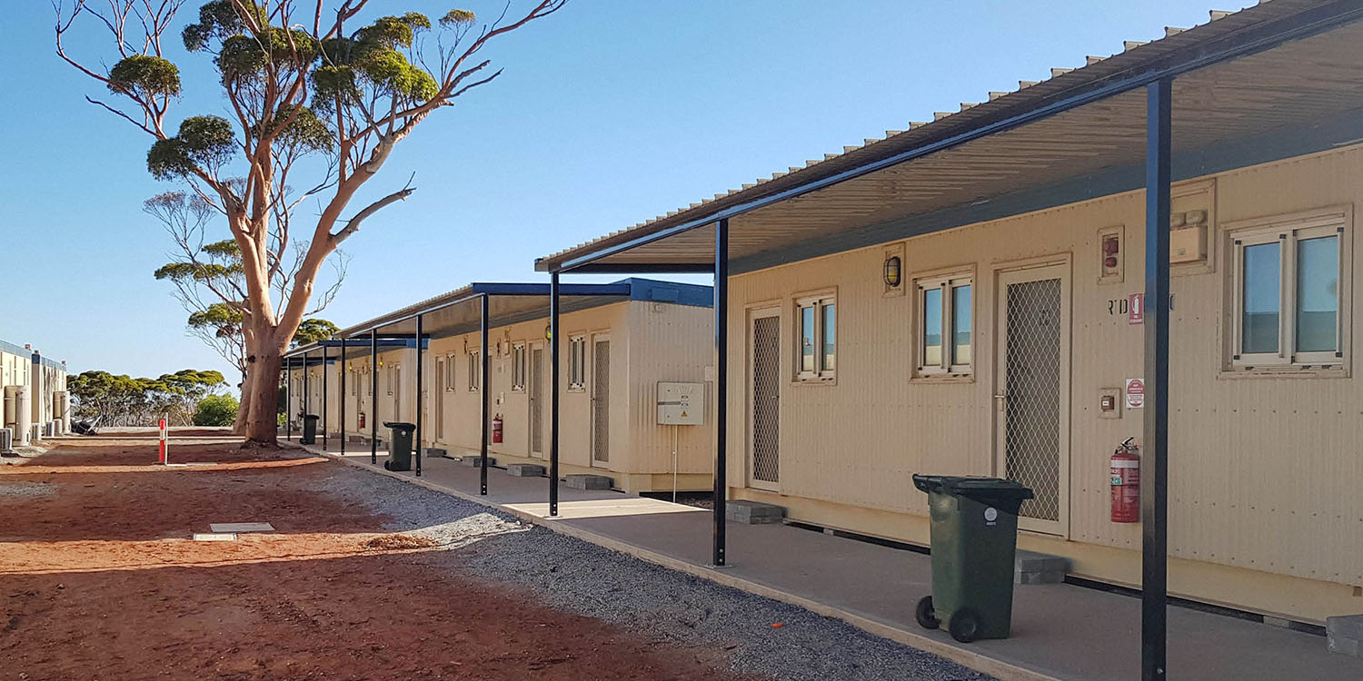Hallway of a mining camp in Western Australia shaded by a large tree and surrounded by red dirt.
