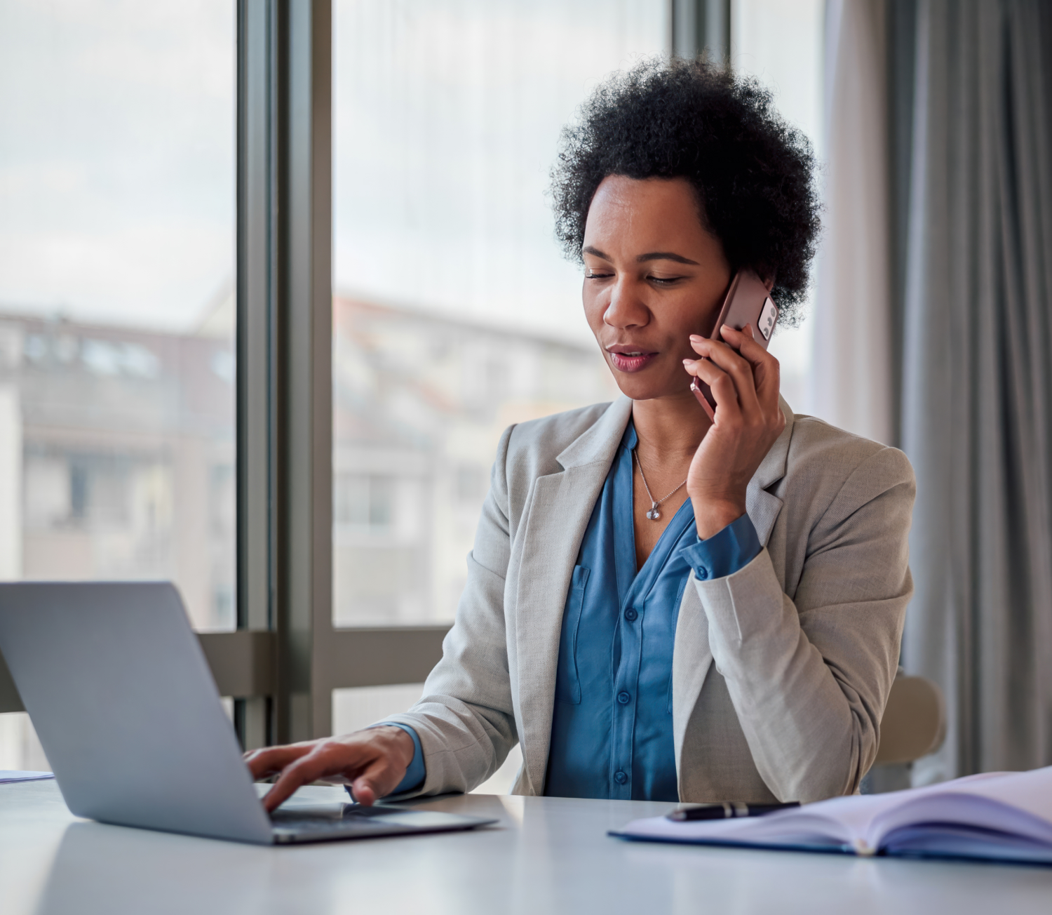 Woman takes call and types at laptop with large window overlooking city behind her.