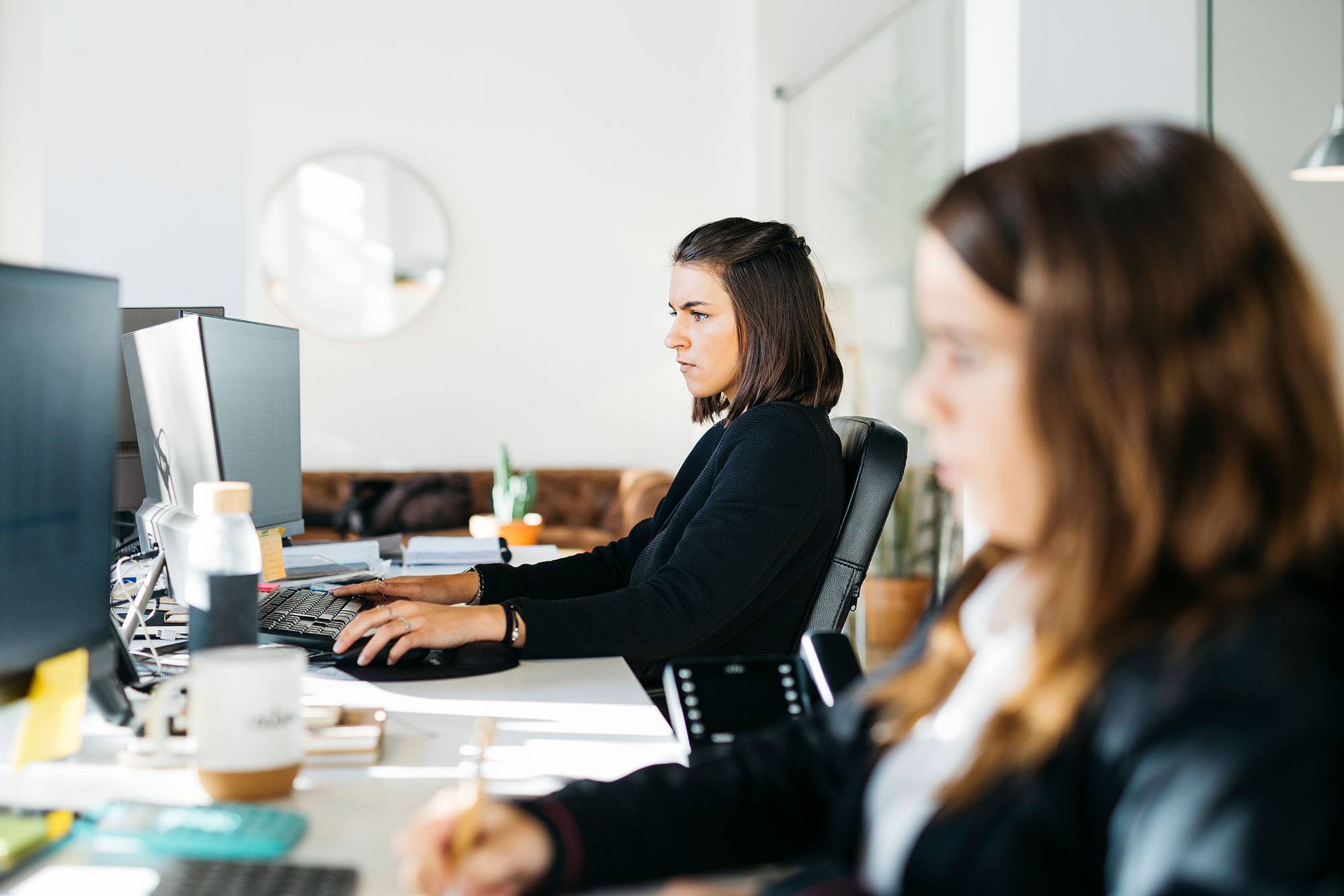Two women in corporate setting sit at a long desk and type at computers.